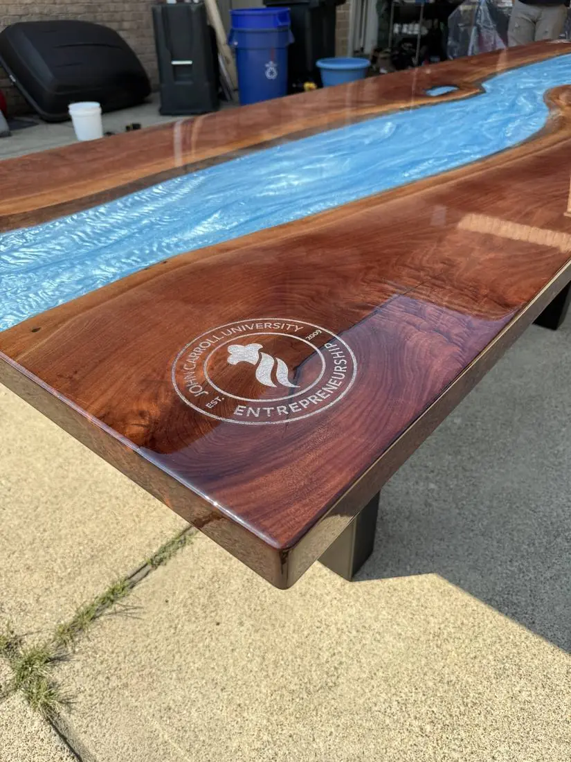Close-up of a glossy wood table corner with a silver engraved circular seal beside a blue epoxy river
