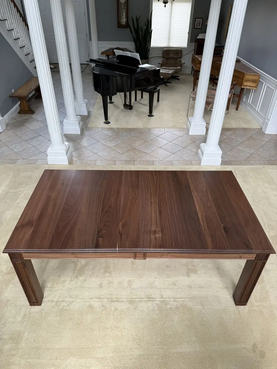 Dark wood dining table seen from above, with a shaped edge and paneled legs, in a hall with white columns and a piano