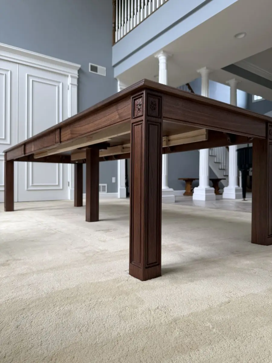 Low view of a dark wood table corner with a paneled leg and small carved flower details at the top
